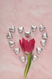 Heart made with delicious chocolate candies and beautiful tulip on pink table, flat lay