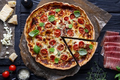 Photo of Cut delicious homemade quiche with prosciutto and ingredients on black wooden table, flat lay