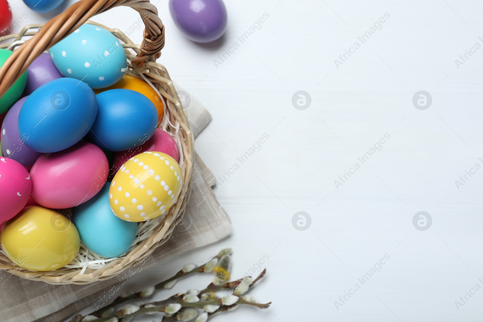 Photo of Colorful Easter eggs in wicker basket and willow branches on white wooden table, flat lay. Space for text