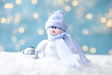 Photo of Snowman toy and Christmas ball on snow against blurred festive lights, closeup