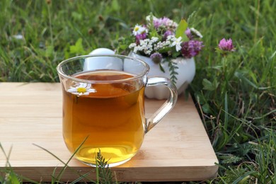 Cup of aromatic herbal tea, pestle and ceramic mortar with different wildflowers on green grass outdoors