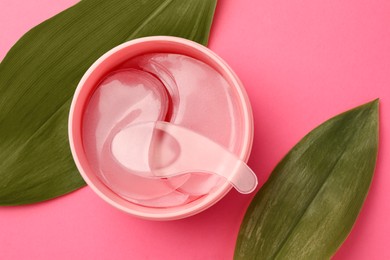 Photo of Jar of under eye patches with spoon and green leaves on pink background, flat lay. Cosmetic product