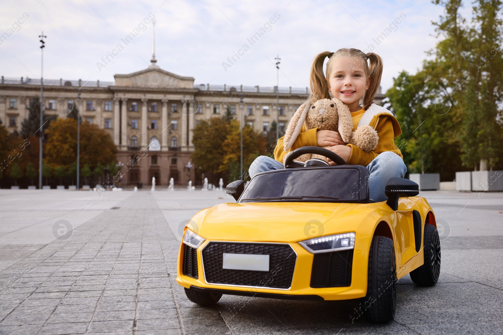 Photo of Cute little girl with toy bunny driving children's car on city street. Space for text