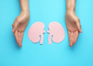 Photo of Woman protecting paper cutout of kidneys on light blue background, top view