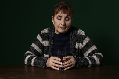 Poor senior woman with glass of water sitting at table against color background