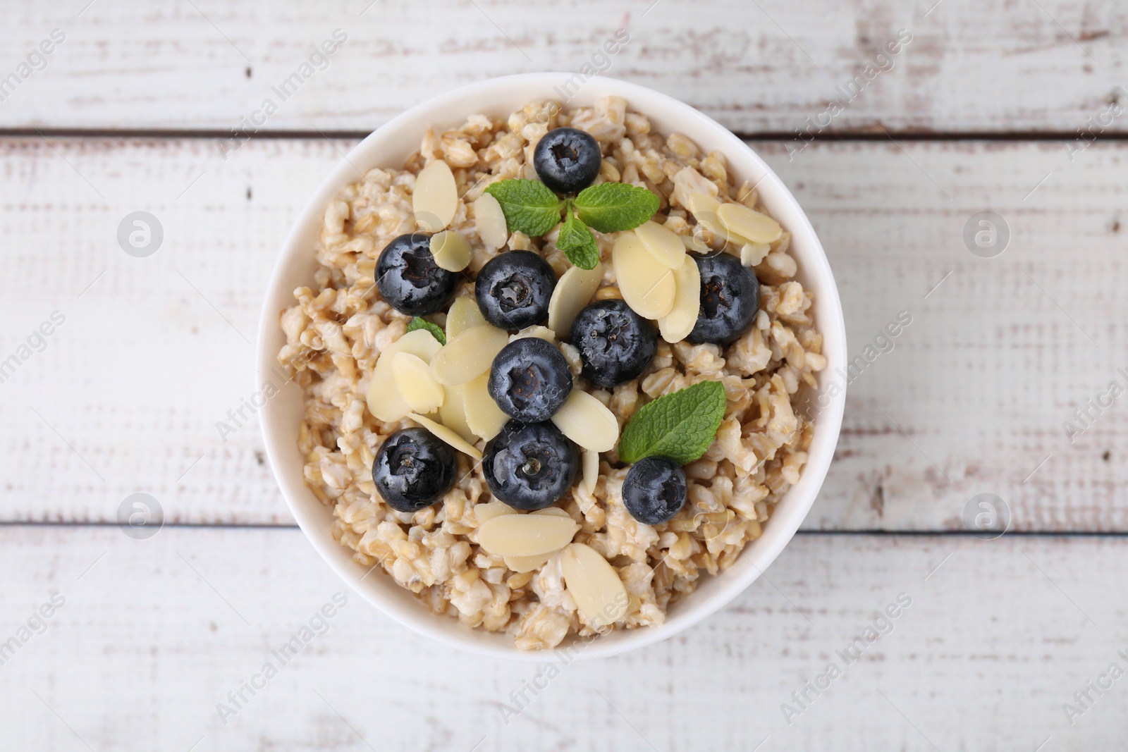 Photo of Tasty oatmeal with blueberries, mint and almond petals in bowl on white wooden table, top view