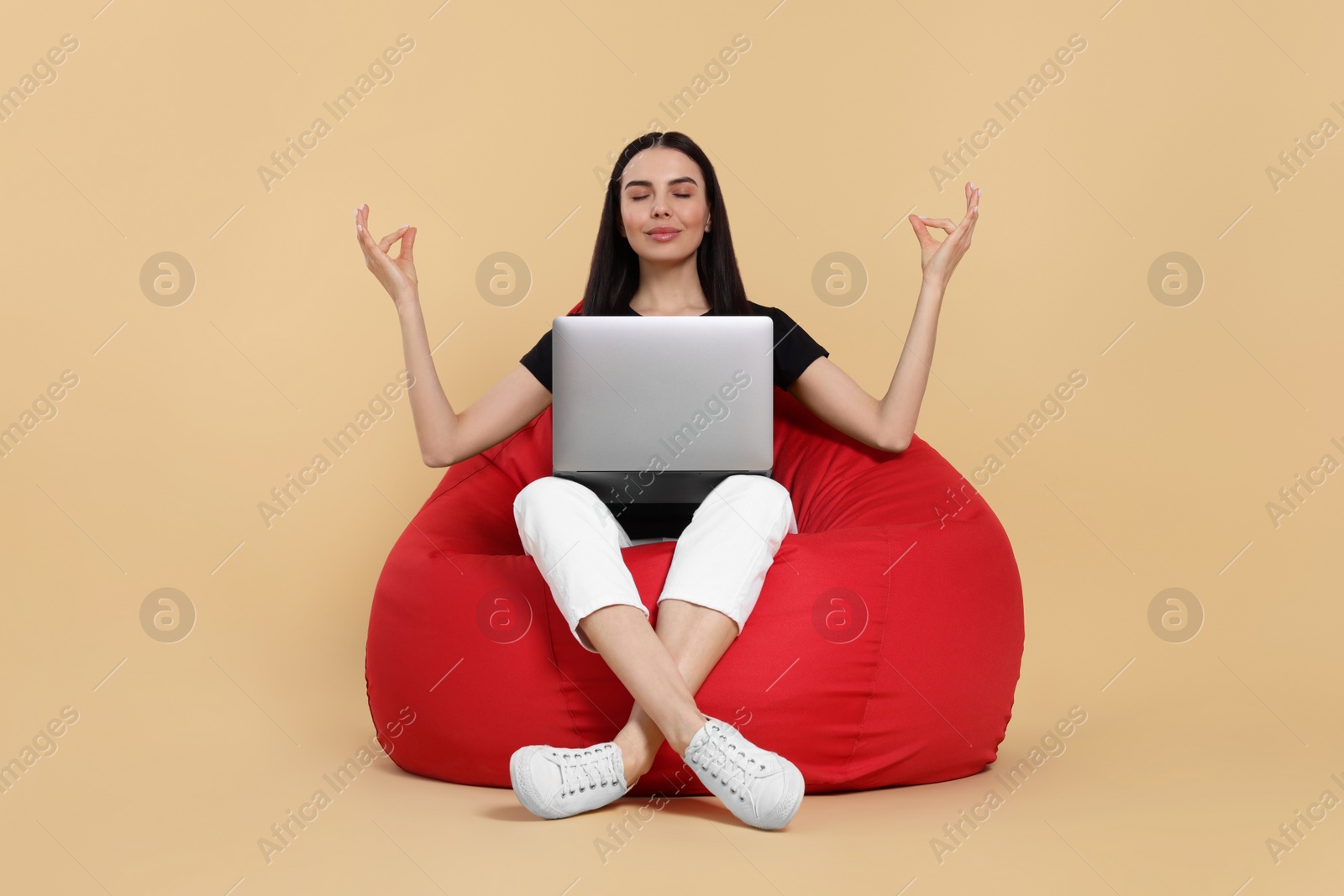 Photo of Beautiful woman with laptop sitting on beanbag chair and meditating against beige background