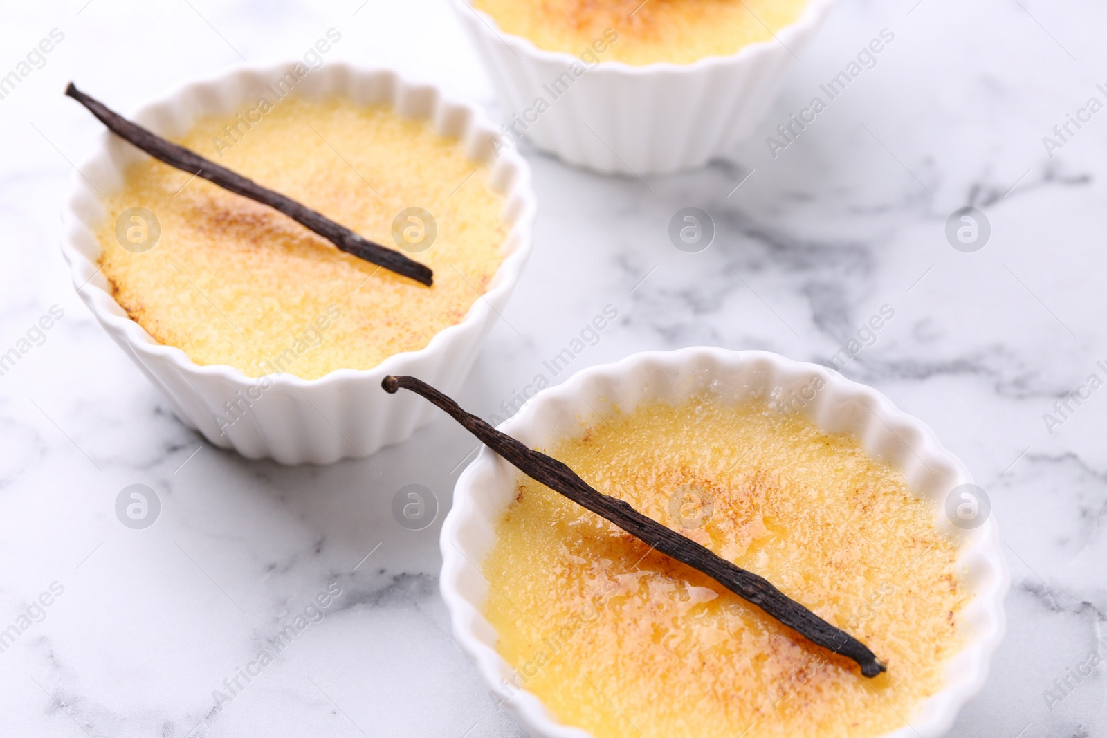 Photo of Delicious creme brulee in bowls and vanilla pods on white marble table, closeup