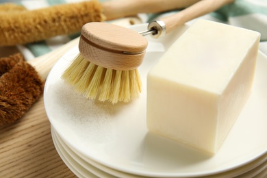 Photo of Cleaning brush, soap bar and plates on wooden table, closeup. Dish washing supplies