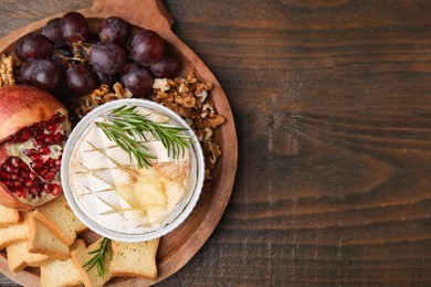 Photo of Board with tasty baked camembert, croutons, grapes, walnuts and pomegranate on wooden table, top view. Space for text