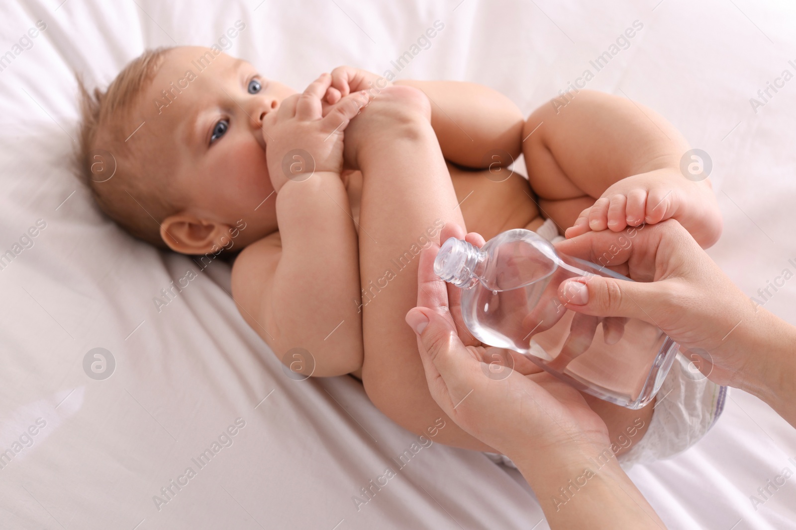Photo of Mother with bottle of massage oil near baby on bed, closeup