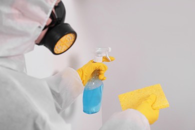 Photo of Woman in protective suit cleaning mold with sprayer and rag on wall indoors, closeup