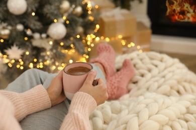 Woman with cup of hot drink resting near fireplace  in living room decorated for Christmas, closeup