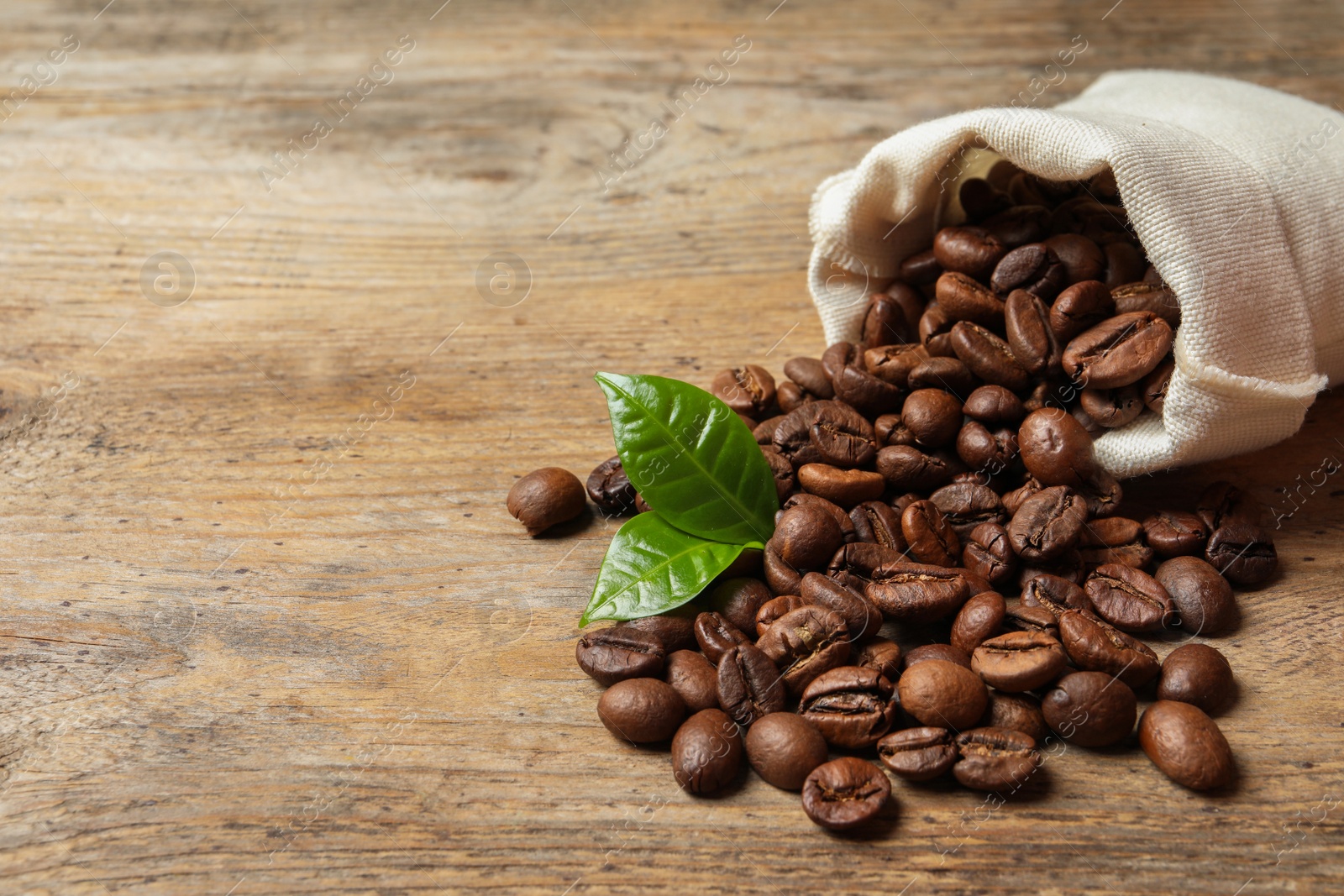 Photo of Bag of coffee beans and fresh green leaves on wooden table, space for text