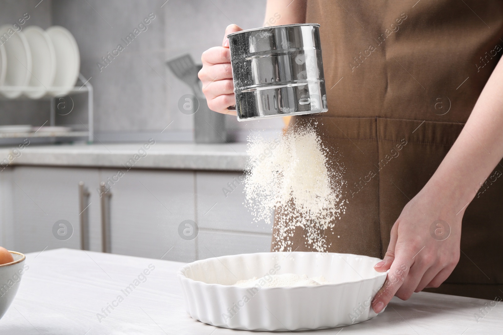 Photo of Woman sieving flour into baking dish at table in kitchen, closeup