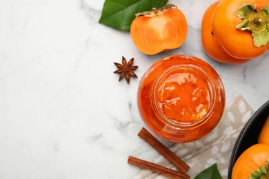 Jar of tasty persimmon jam and ingredients on white marble table, flat lay. Space for text