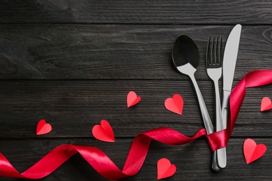 Photo of Cutlery set and red ribbon on black wooden background, flat lay with space for text. Valentine's Day dinner
