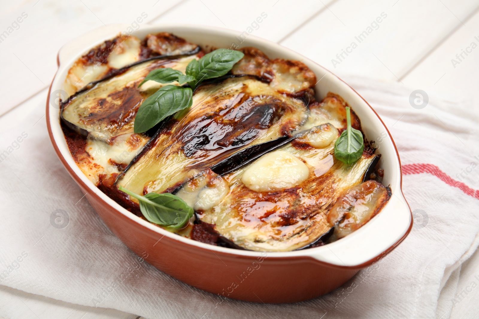 Photo of Delicious eggplant lasagna in baking dish and napkin on white wooden table, closeup