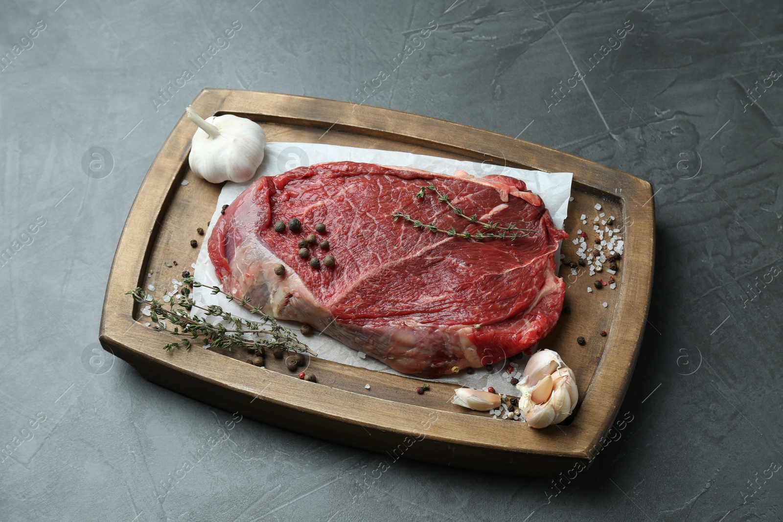 Photo of Wooden tray with piece of raw meat, garlic and thyme on grey table