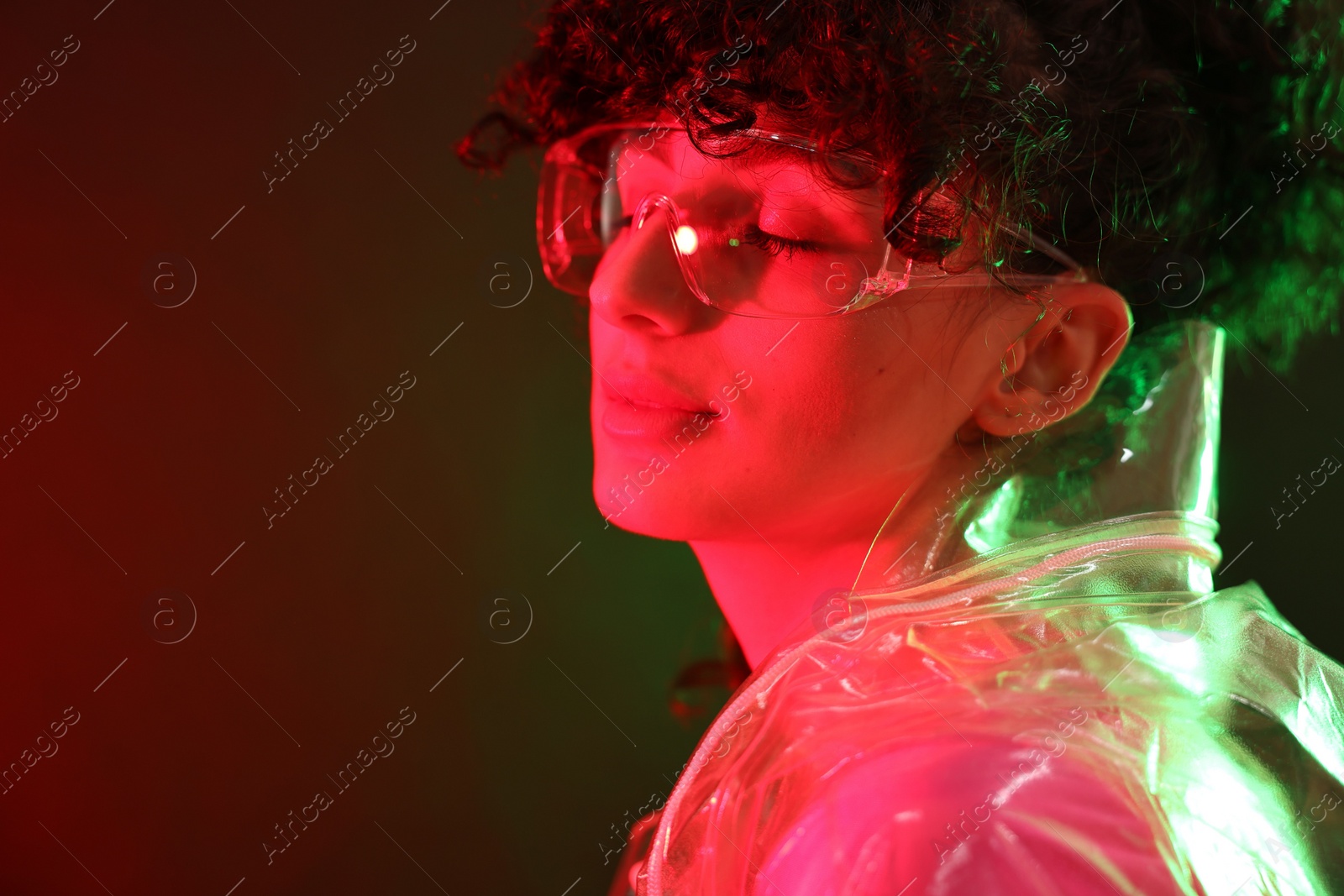 Photo of Beautiful young woman in transparent coat and sunglasses posing on color background in neon lights. Space for text