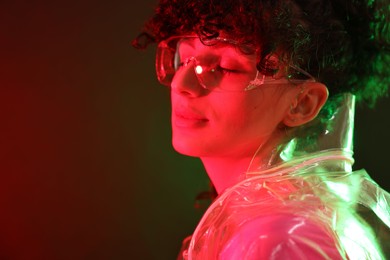 Photo of Beautiful young woman in transparent coat and sunglasses posing on color background in neon lights. Space for text