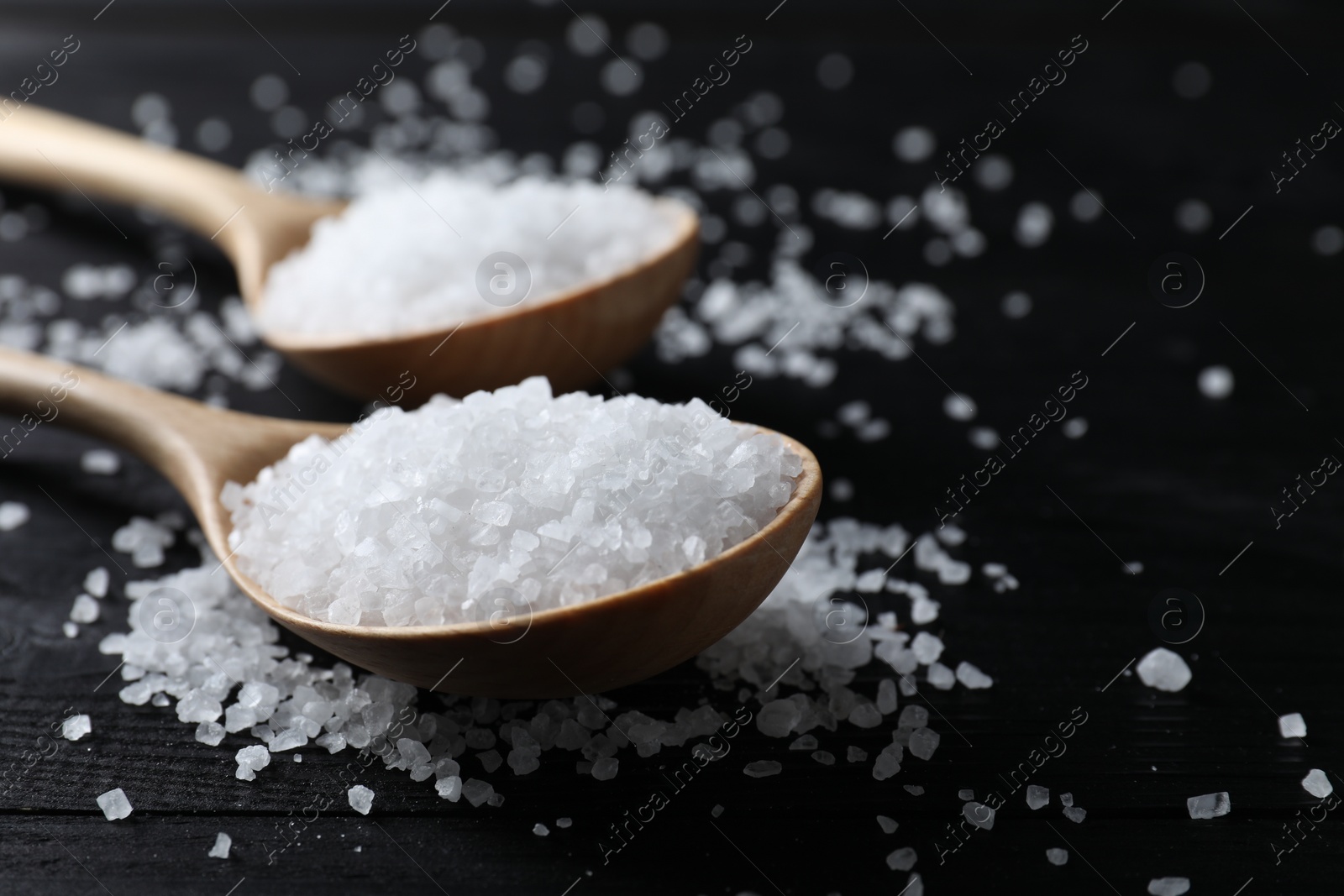 Photo of Organic salt in spoons on black wooden table, closeup. Space for text