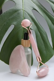 Photo of Gua sha stone, face roller, bottle of serum and monstera leaf on light background