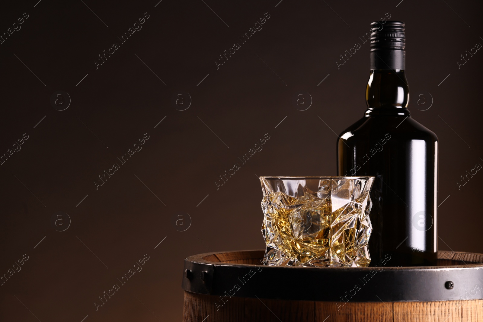 Photo of Whiskey with ice cubes in glass and bottle on wooden barrel against dark background, space for text