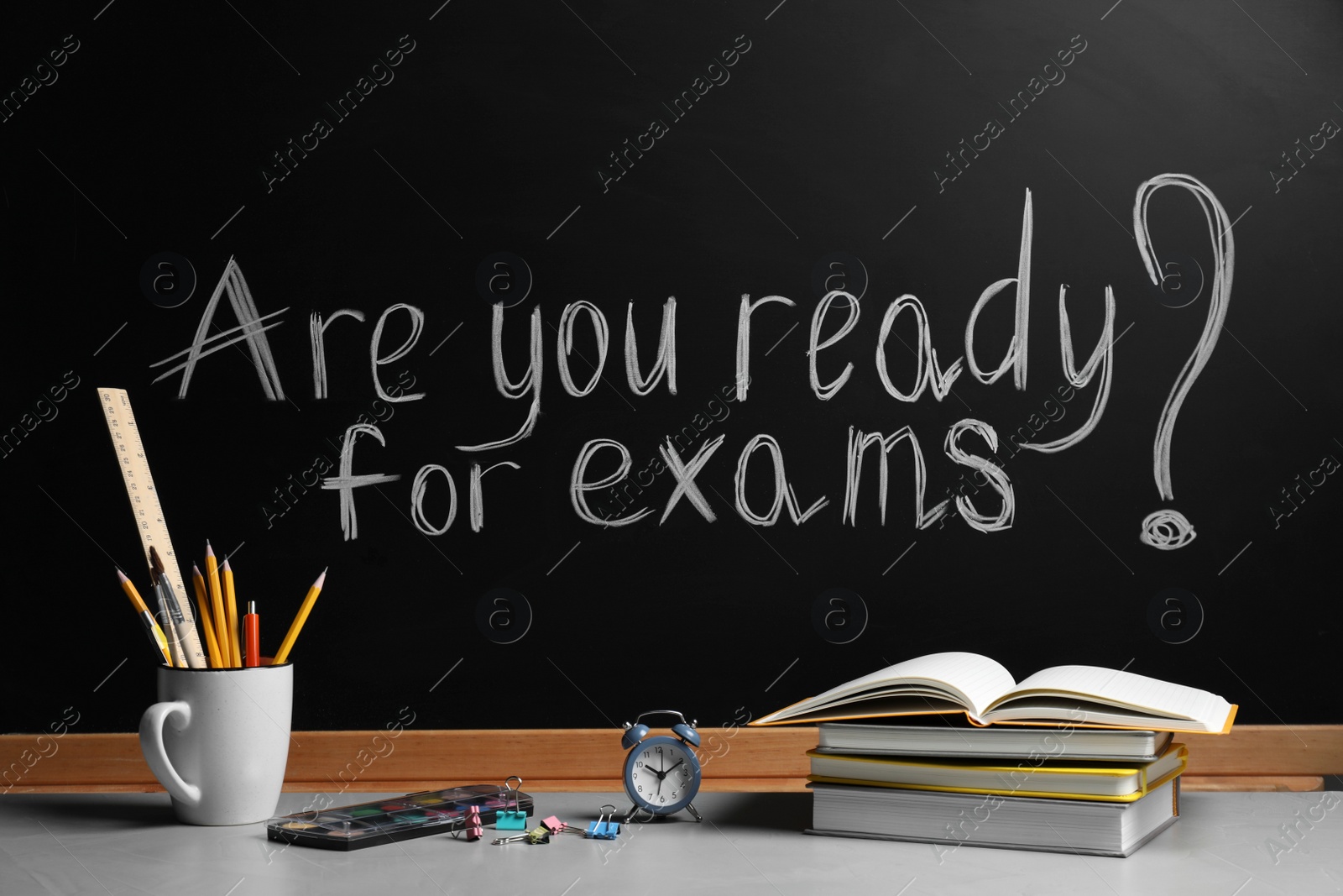 Photo of Different stationery and alarm clock on light table near blackboard with phrase Are You Ready For Exams