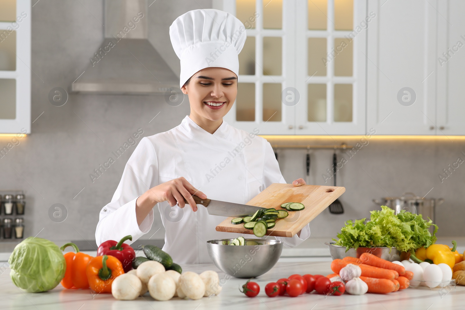 Photo of Professional chef putting cut cucumber into metal bowl at white marble table in kitchen