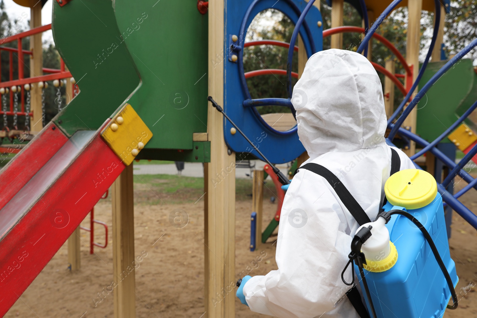 Photo of Woman wearing chemical protective suit with disinfectant sprayer on playground. Preventive measure during coronavirus pandemic