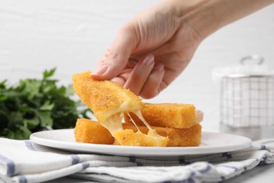 Photo of Woman taking tasty fried mozzarella stick from plate at table, closeup