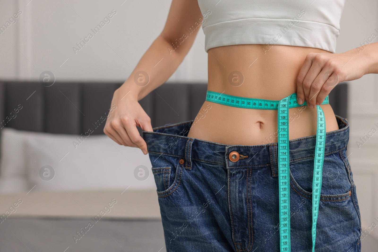 Photo of Closeup view of slim woman wearing big jeans and measuring waist with tape in room, space for text. Weight loss