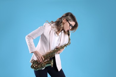 Beautiful young woman in elegant outfit playing saxophone on light blue background