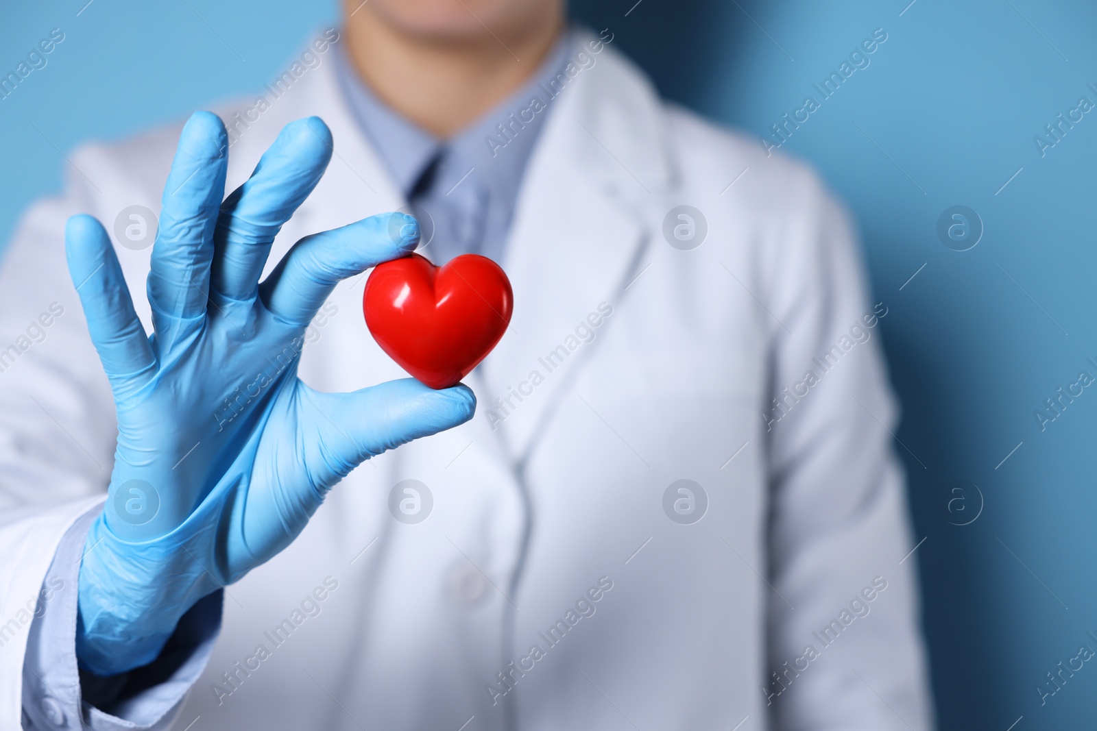 Photo of Doctor wearing medical gloves holding decorative heart on light blue background, closeup