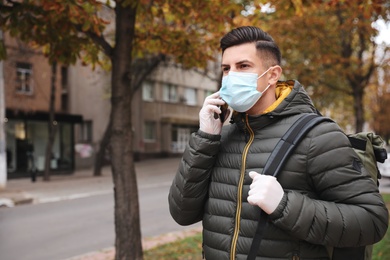 Photo of Man in medical face mask and gloves talking on phone while walking outdoors. Personal protection during COVID-19 pandemic