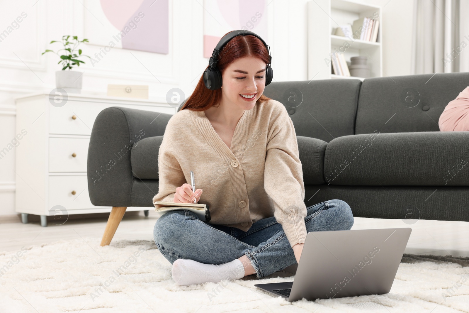 Photo of Happy woman with headphones and notebook using laptop on rug in living room