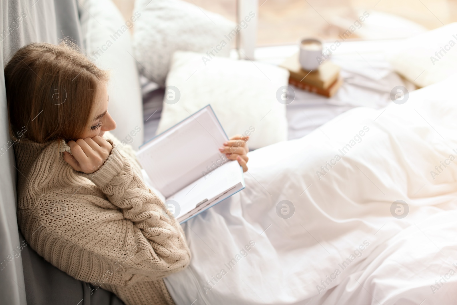 Photo of Beautiful young woman in knitted sweater sitting and reading book near window at home. Winter atmosphere