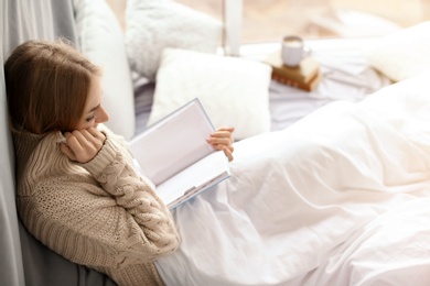 Photo of Beautiful young woman in knitted sweater sitting and reading book near window at home. Winter atmosphere