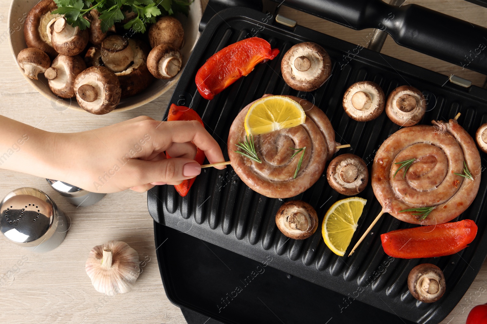 Photo of Woman cooking homemade sausages with mushrooms and bell pepper on electric grill at wooden table, top view