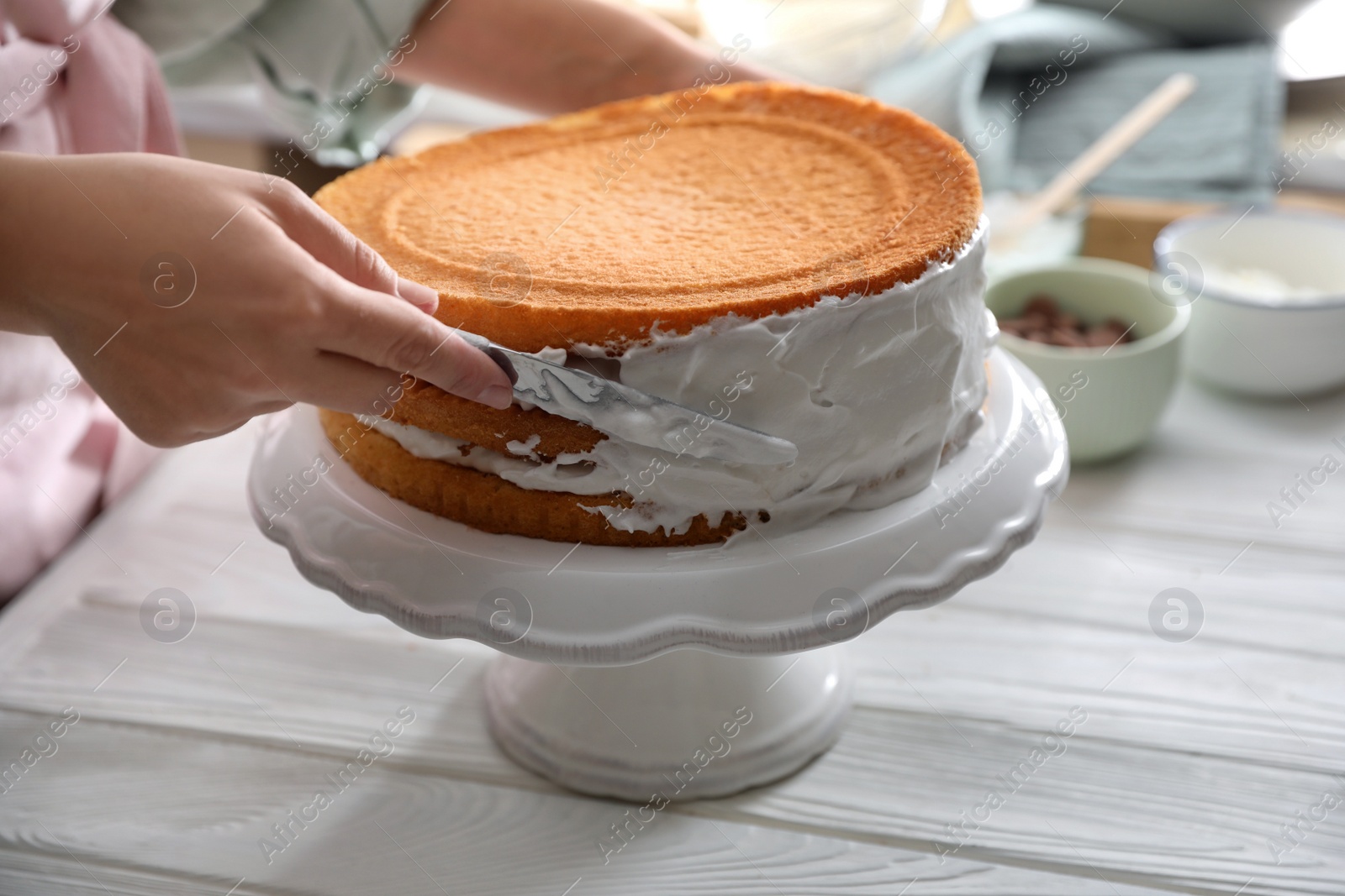 Photo of Woman smearing sides of sponge cake with cream at white wooden table, closeup