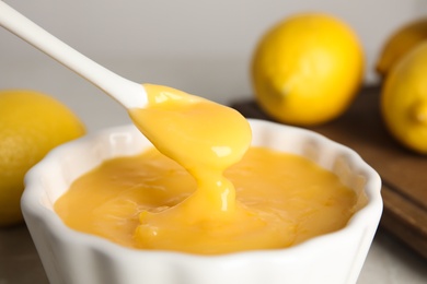 Spoon with delicious lemon curd above bowl on table, closeup