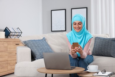 Muslim woman using smartphone near laptop at couch in room. Space for text