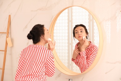 Photo of Young woman cleaning face with cotton pad near mirror in bathroom