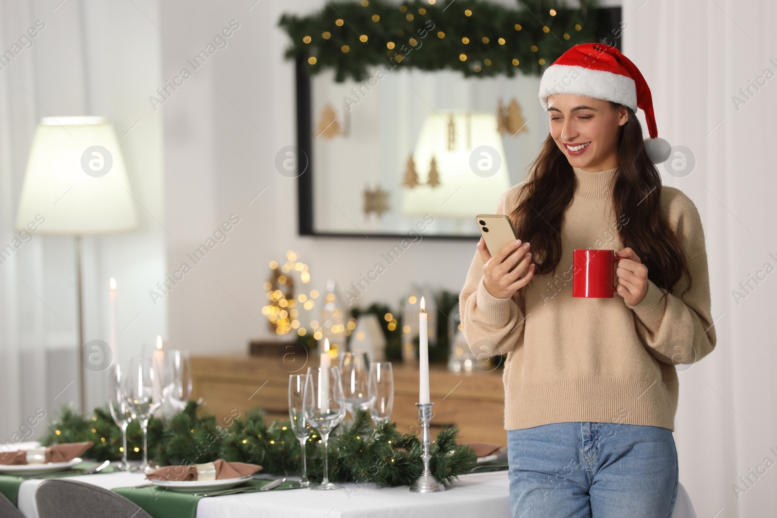 Photo of Smiling woman in Santa hat holding cup and smartphone near table with Christmas setting at home. Space for text