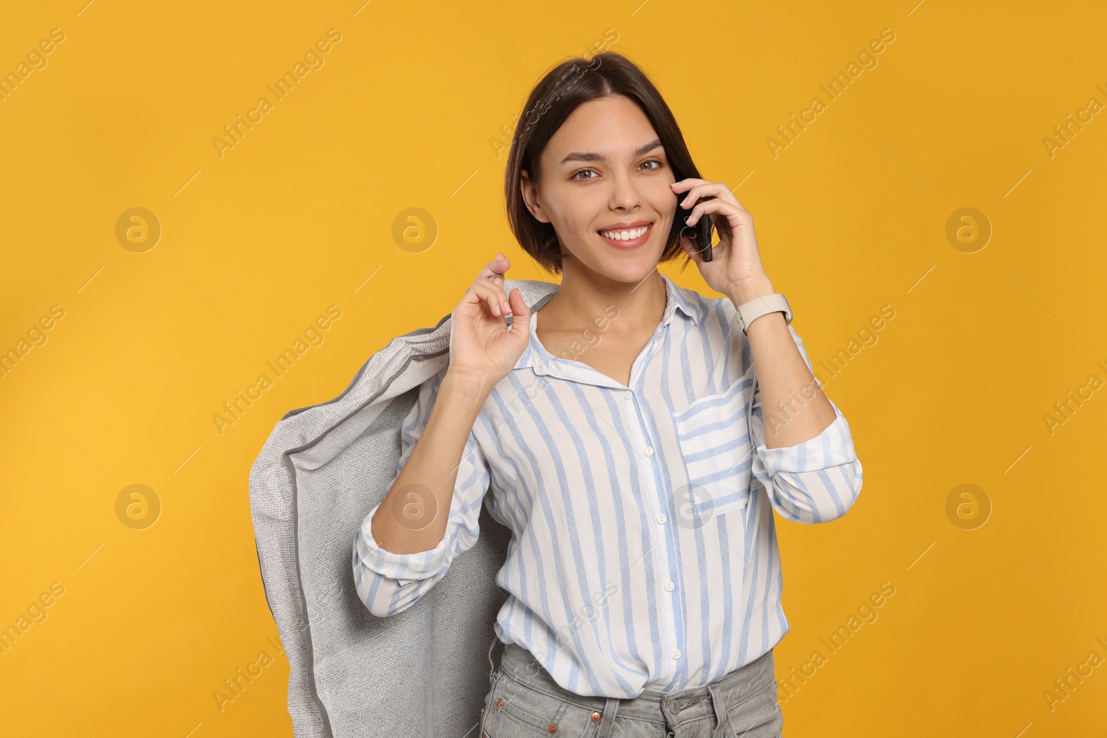 Photo of Woman holding garment cover with clothes while talking on phone against yellow background. Dry-cleaning service