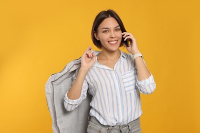 Photo of Woman holding garment cover with clothes while talking on phone against yellow background. Dry-cleaning service