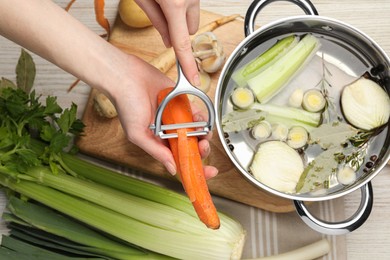 Photo of Cooking tasty bouillon. Woman peeling carrot at white wooden table, top view