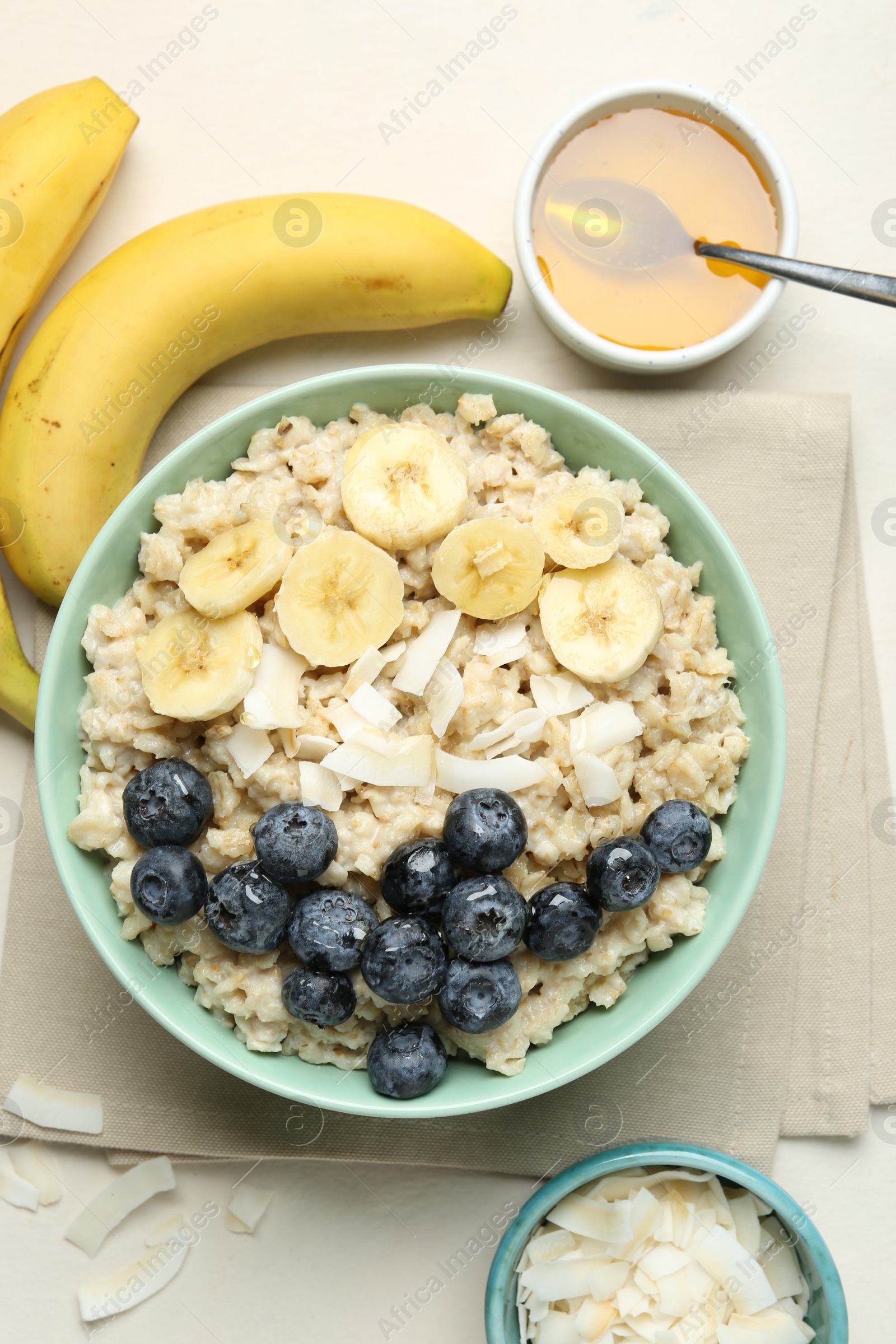 Photo of Tasty oatmeal with banana, blueberries, coconut flakes and honey served in bowl on beige table, flat lay
