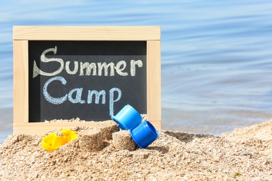 Photo of Little blackboard with text SUMMER CAMP and toys on sand near sea